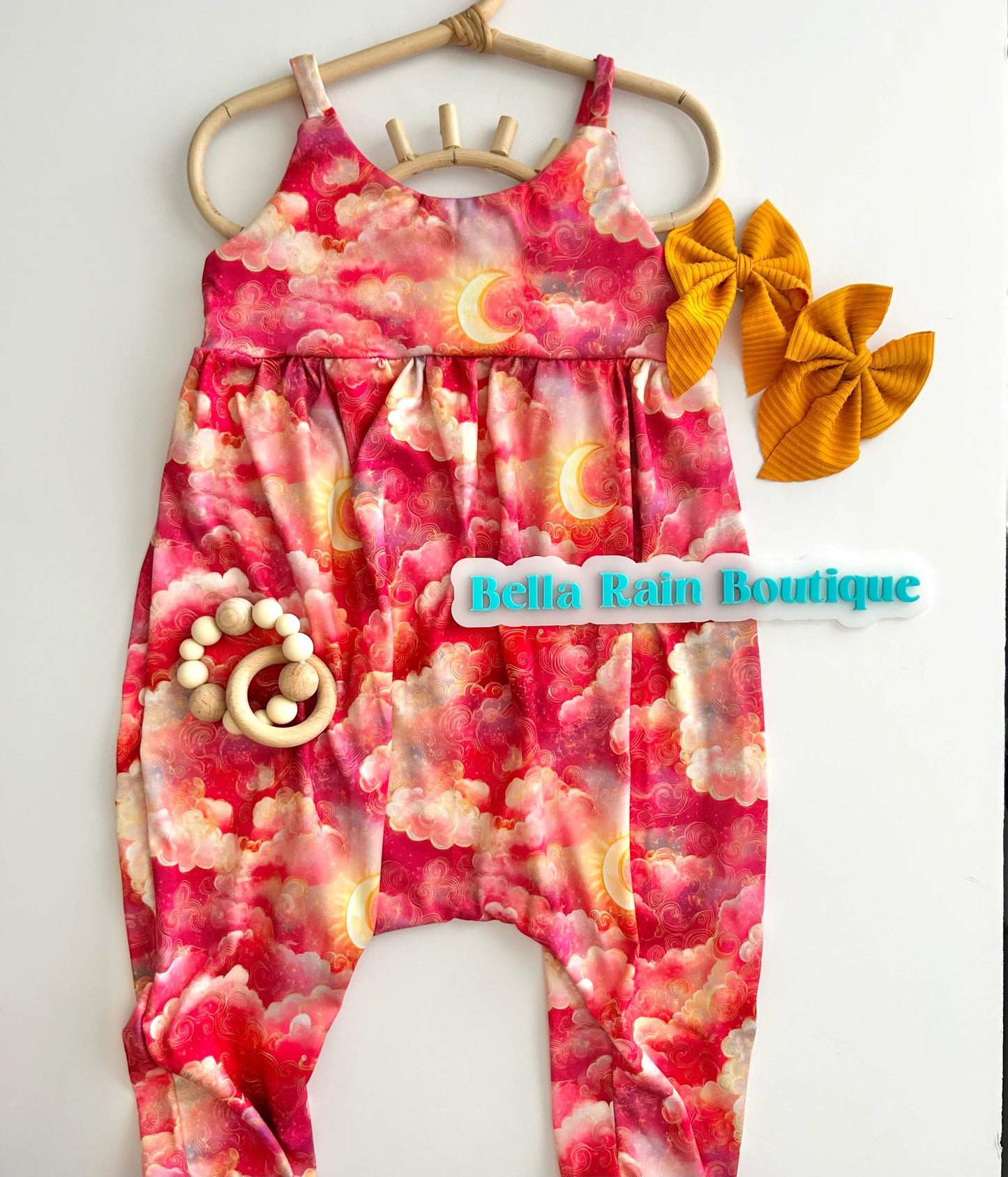 A pink strappy romper with a sky print, displayed next to a wooden hoop and a yellow bow.