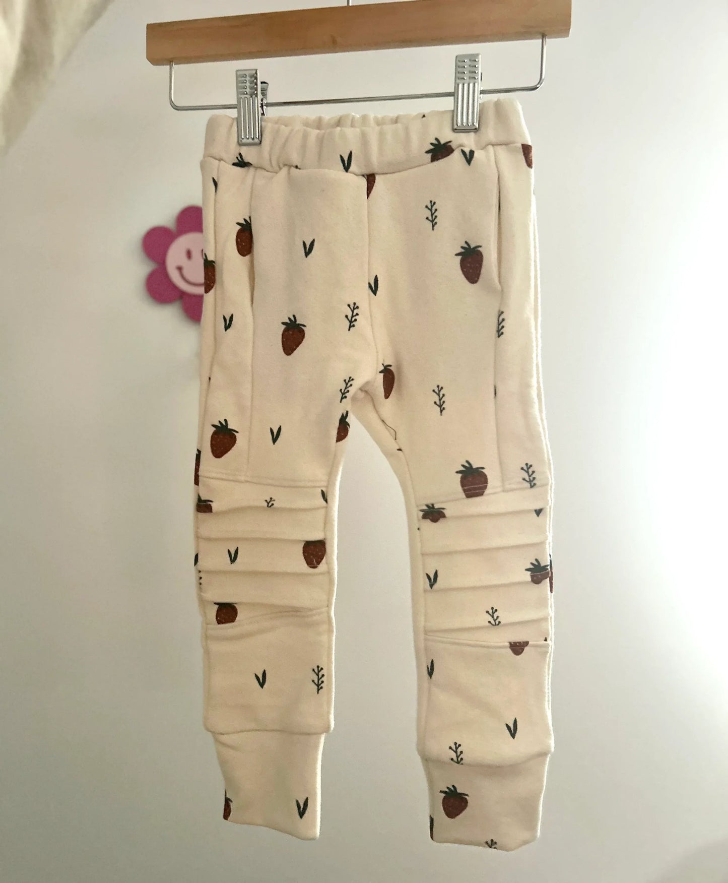 Beige baby pocket jogger pants with strawberry pattern hanging on a rack against a white background. Handmade kids apparel for kids in sizes Nb-10.