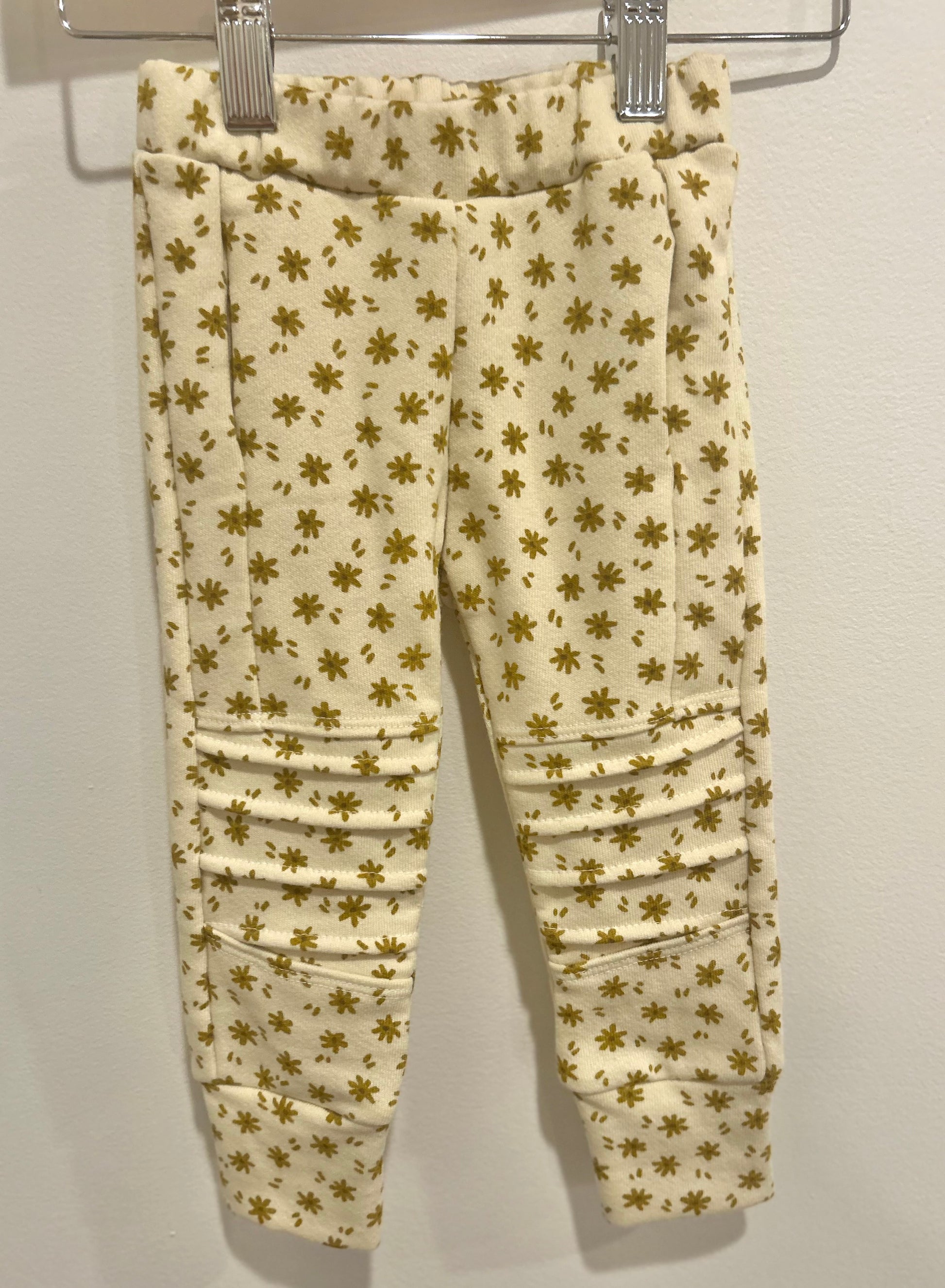 Beige pants with gold floral pattern hanging on a rack against a white background from Bella Rain Boutique. A handmade kids clothing brand