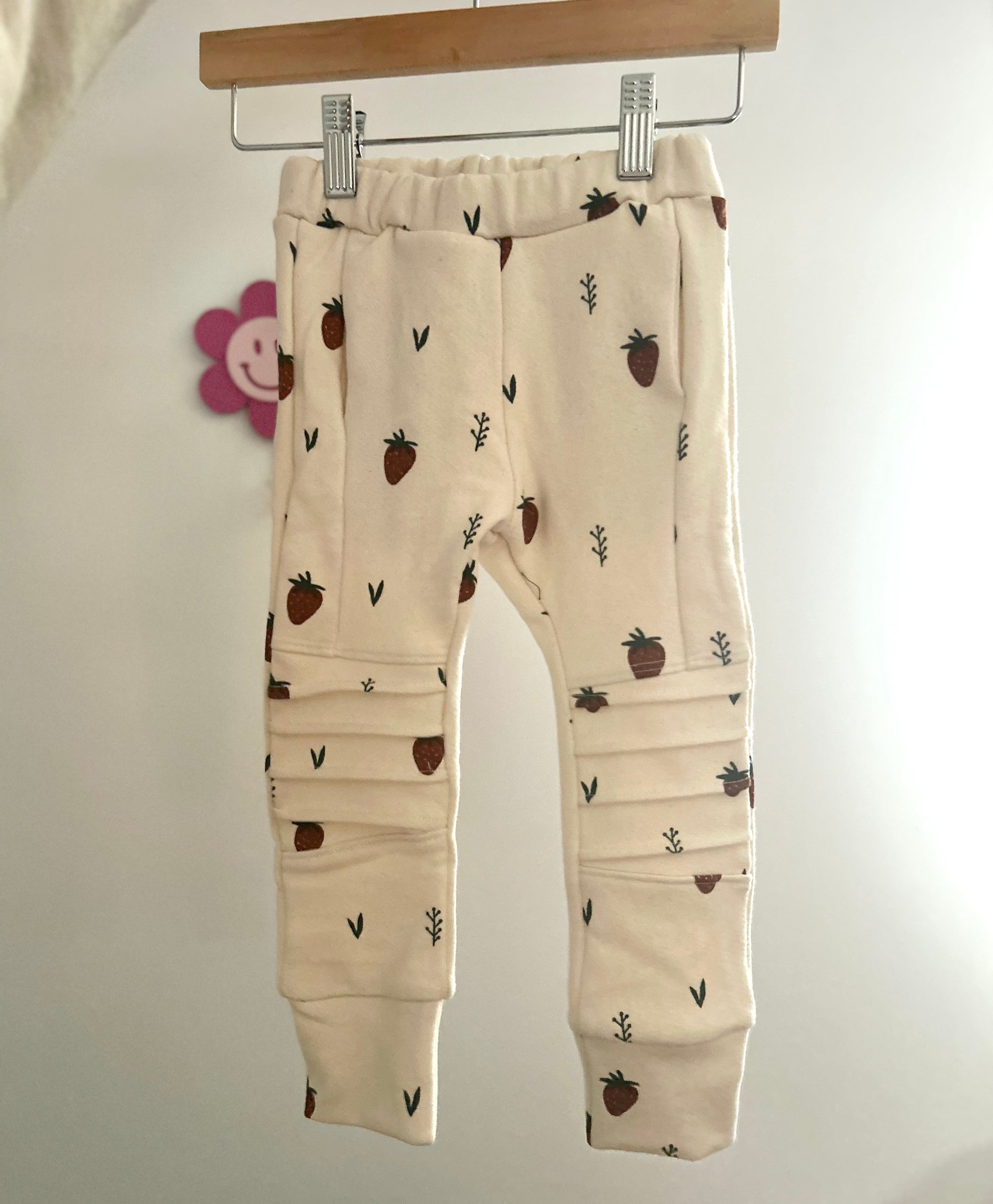 Beige baby pocket jogger pants with strawberry pattern hanging on a rack against a white background. Handmade kids apparel for kids in sizes Nb-10.