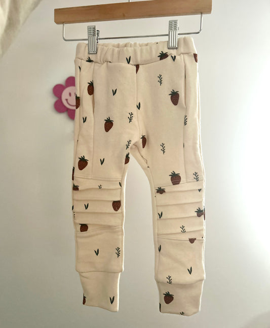 Beige baby pocket jogger pants with strawberry pattern hanging on a rack against a white background. Handmade kids apparel for kids in sizes Nb-10.